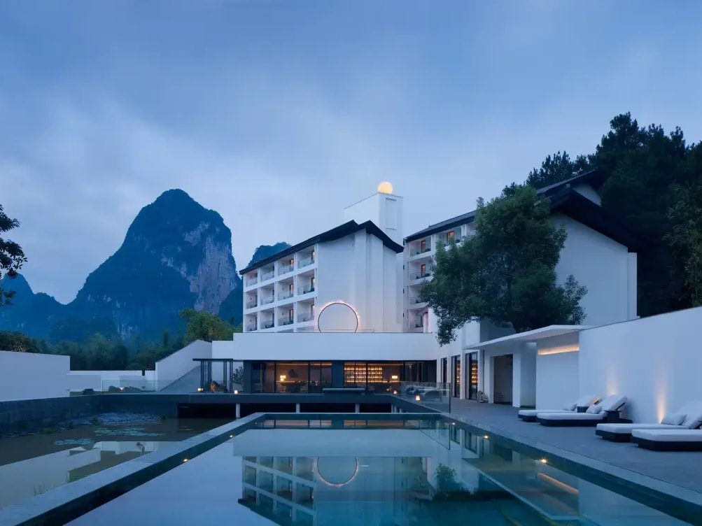 White low-rise hotel blocks at dusk mirrored in a long still pool, karst peak behind