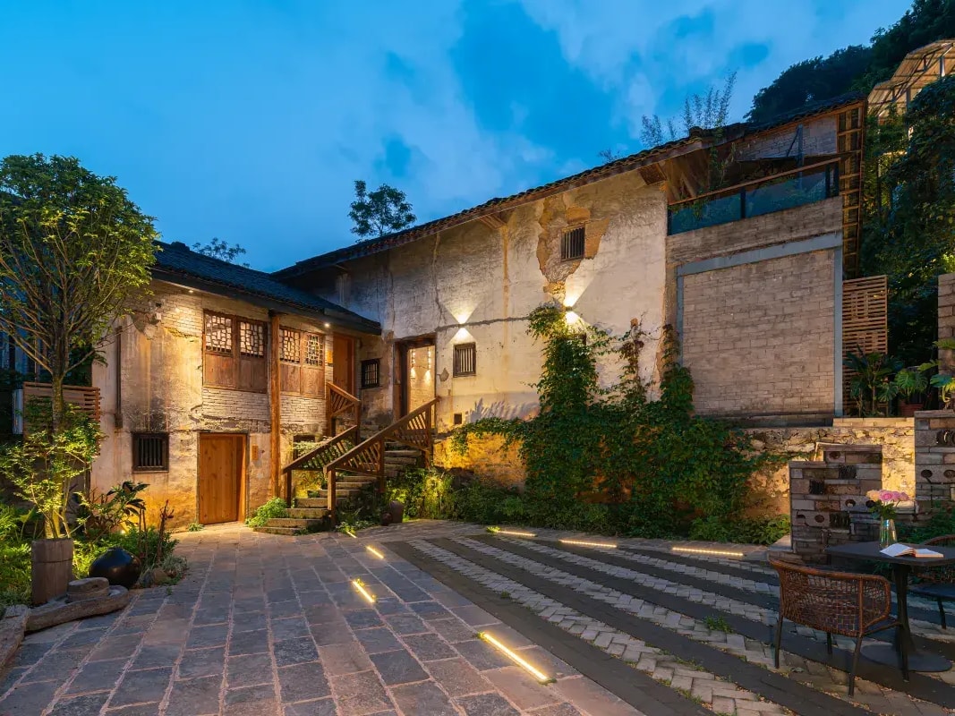 Restored stone farmhouse lit at dusk above a paved forecourt with a table and chairs
