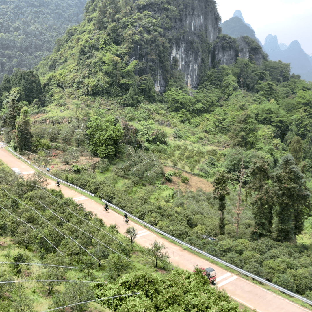 Aerial view of riders on a rural road below a limestone cliff, a three-wheeler ahead of them