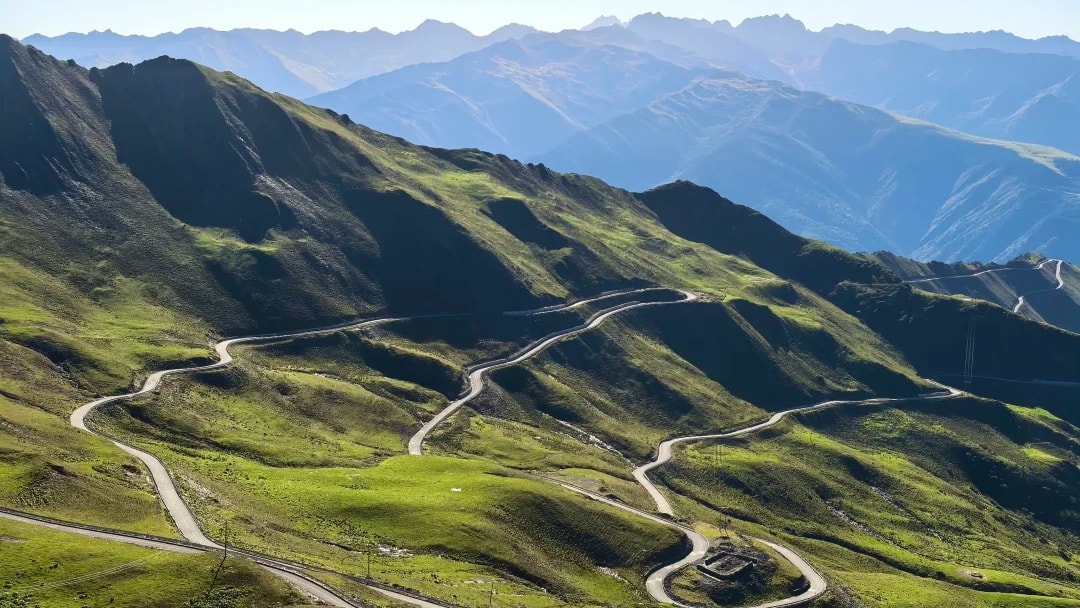 Switchback mountain road winding across green alpine slopes above small tarns.