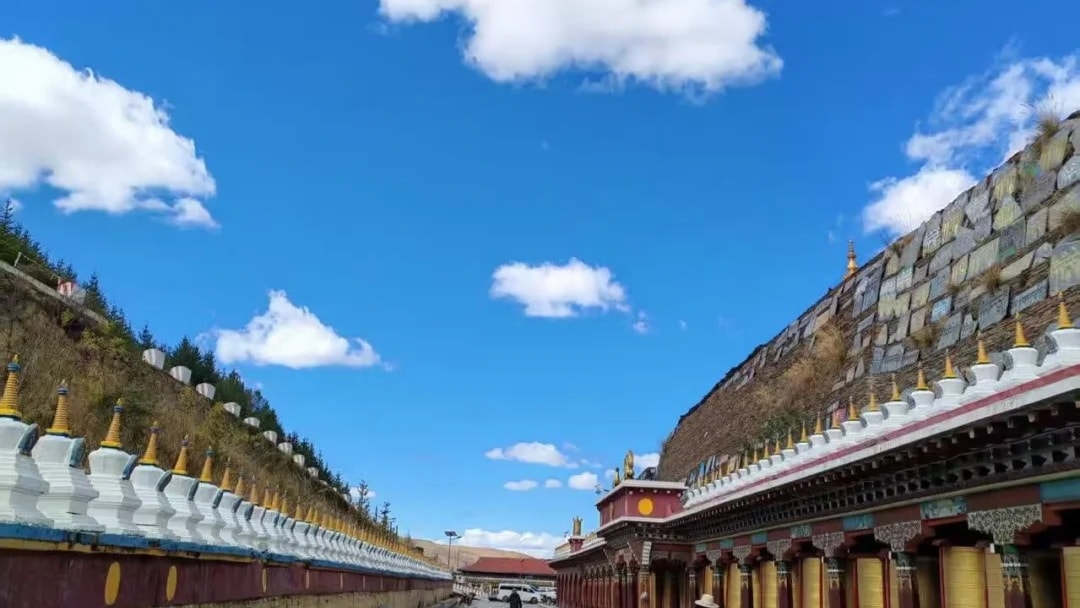 A long corridor of Tibetan prayer wheels running beside a paved plaza under a blue sky.