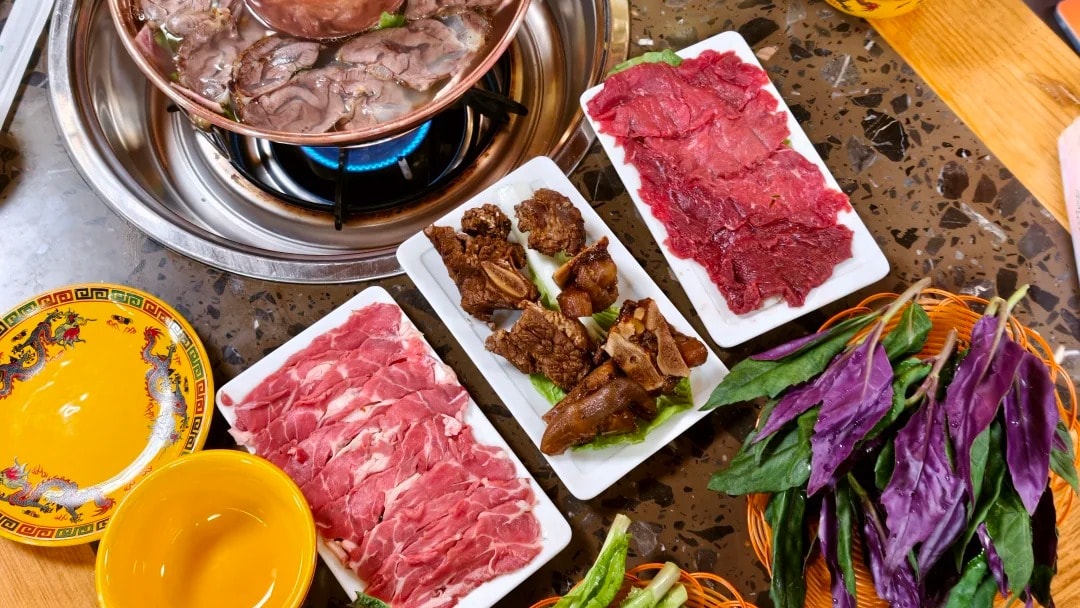 A copper yak hotpot on a wooden table with plates of sliced meat, greens and butter tea.