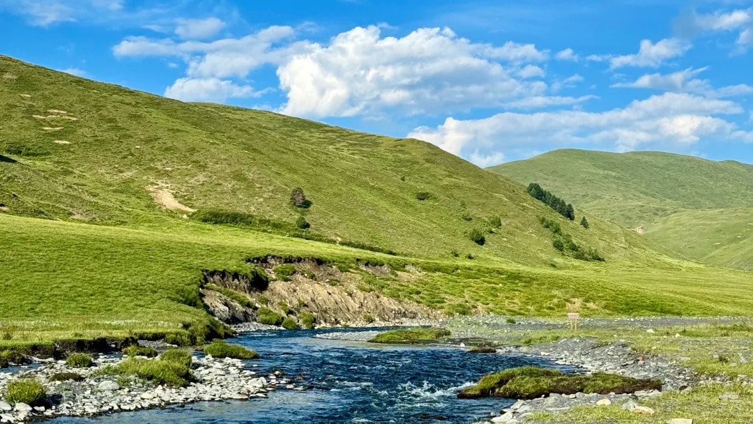 A clear stream winding through green alpine meadow under a bright blue sky on Jiajin Mountain.