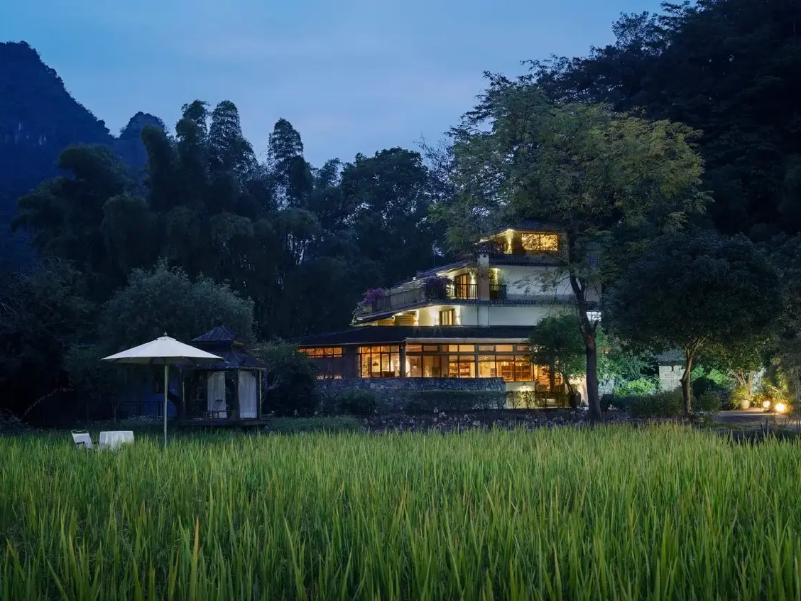 Lodge lit from within at dusk behind a green rice paddy, wooded karst hill above