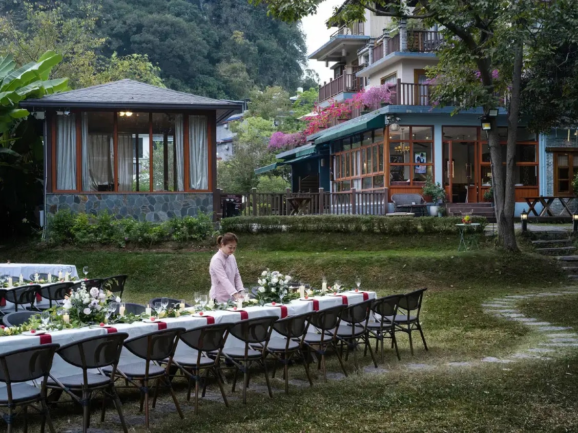 Long table laid with flowers and candles on a lawn, timber and glass guesthouse buildings behind