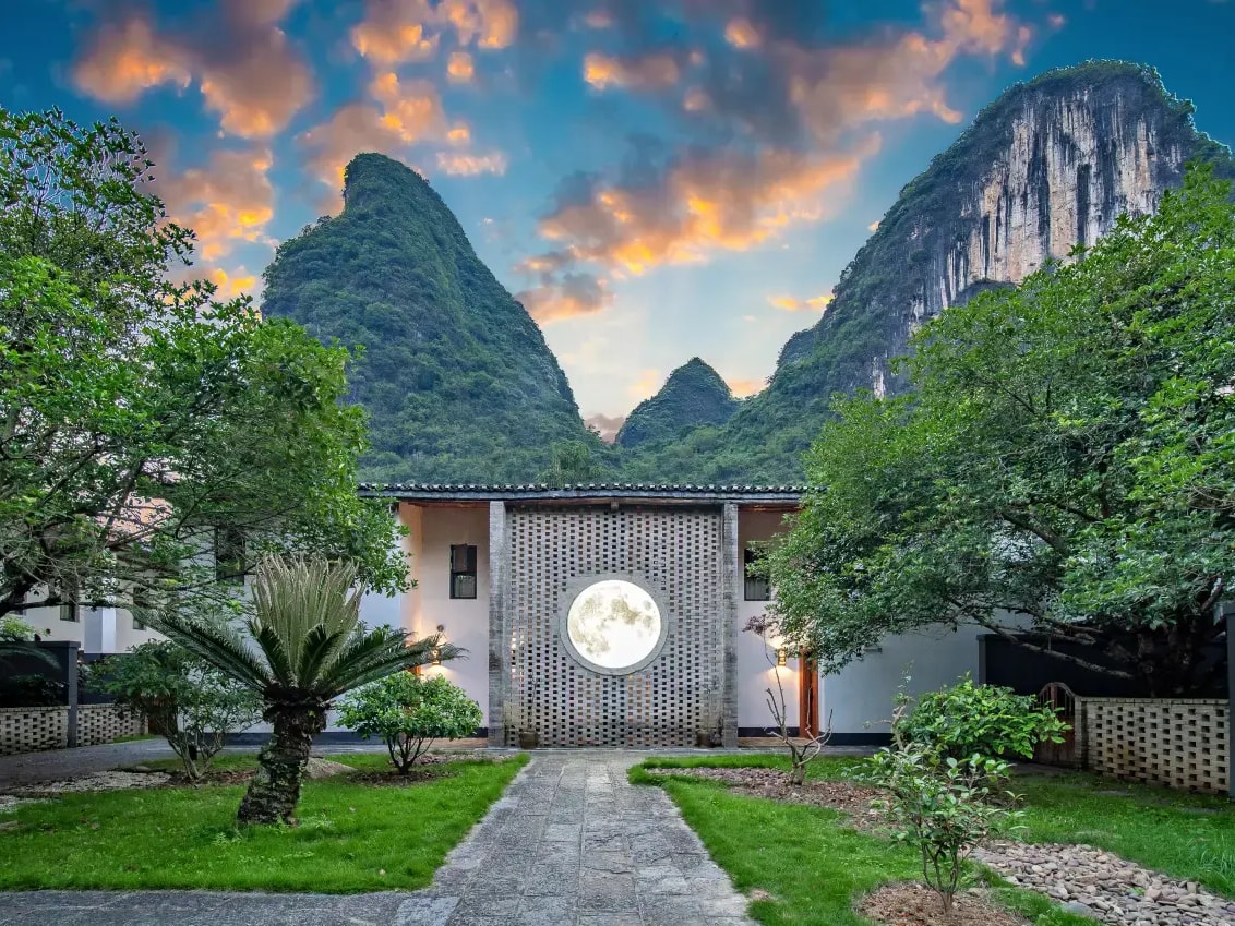 Lawn path to a perforated brick wall holding a glowing moon disc, karst peaks at sunset behind