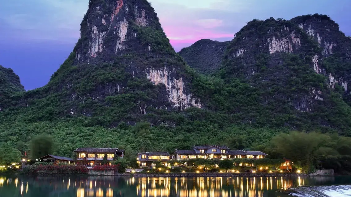 Low-rise lodge lit at dusk on a river bank, karst peaks above and lights reflected in the water