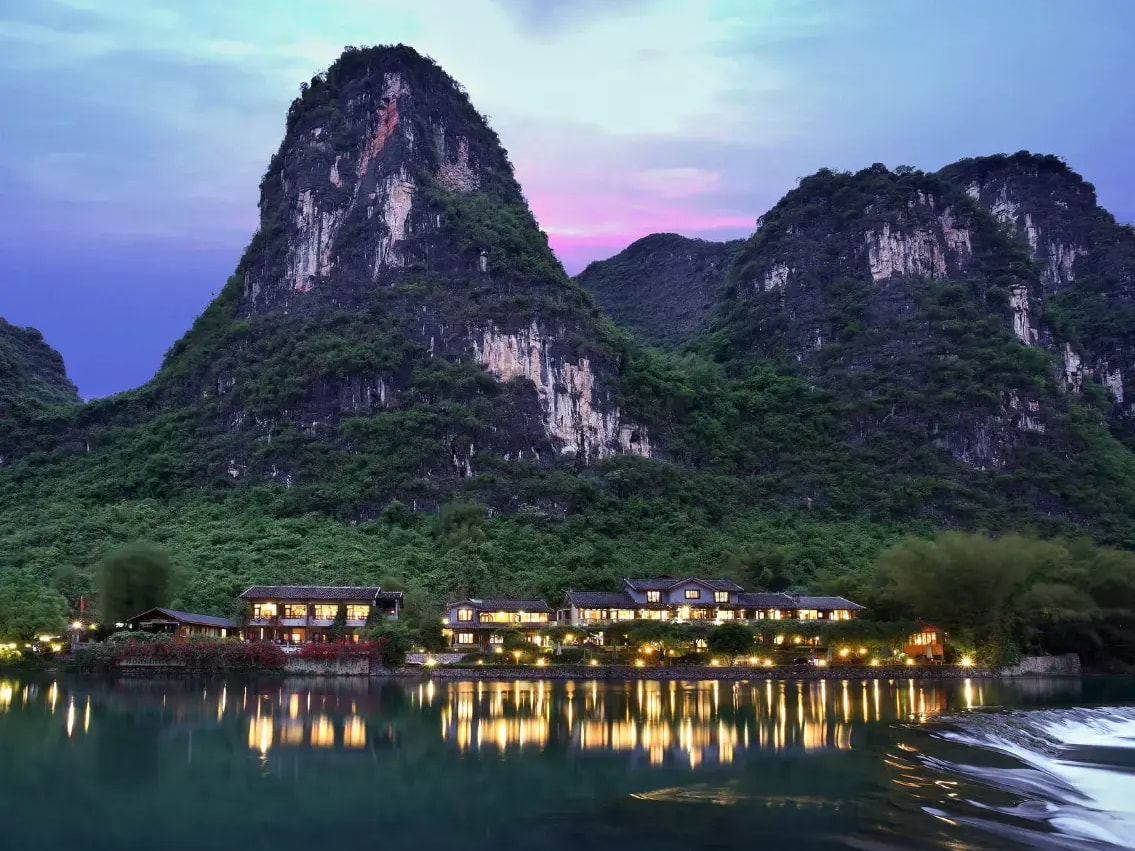 Low-rise lodge lit at dusk on a river bank, karst peaks above and lights reflected in the water