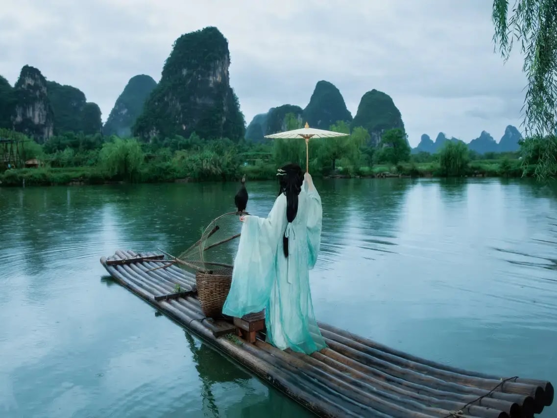 Woman in flowing robes with a paper parasol on a bamboo raft, cormorant beside her, karst peaks behind