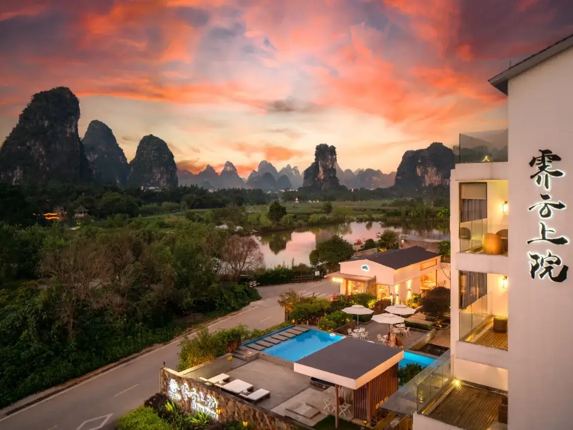 Hotel terrace and lit pool at sunset, river and karst peaks beyond, name carved on the white wall
