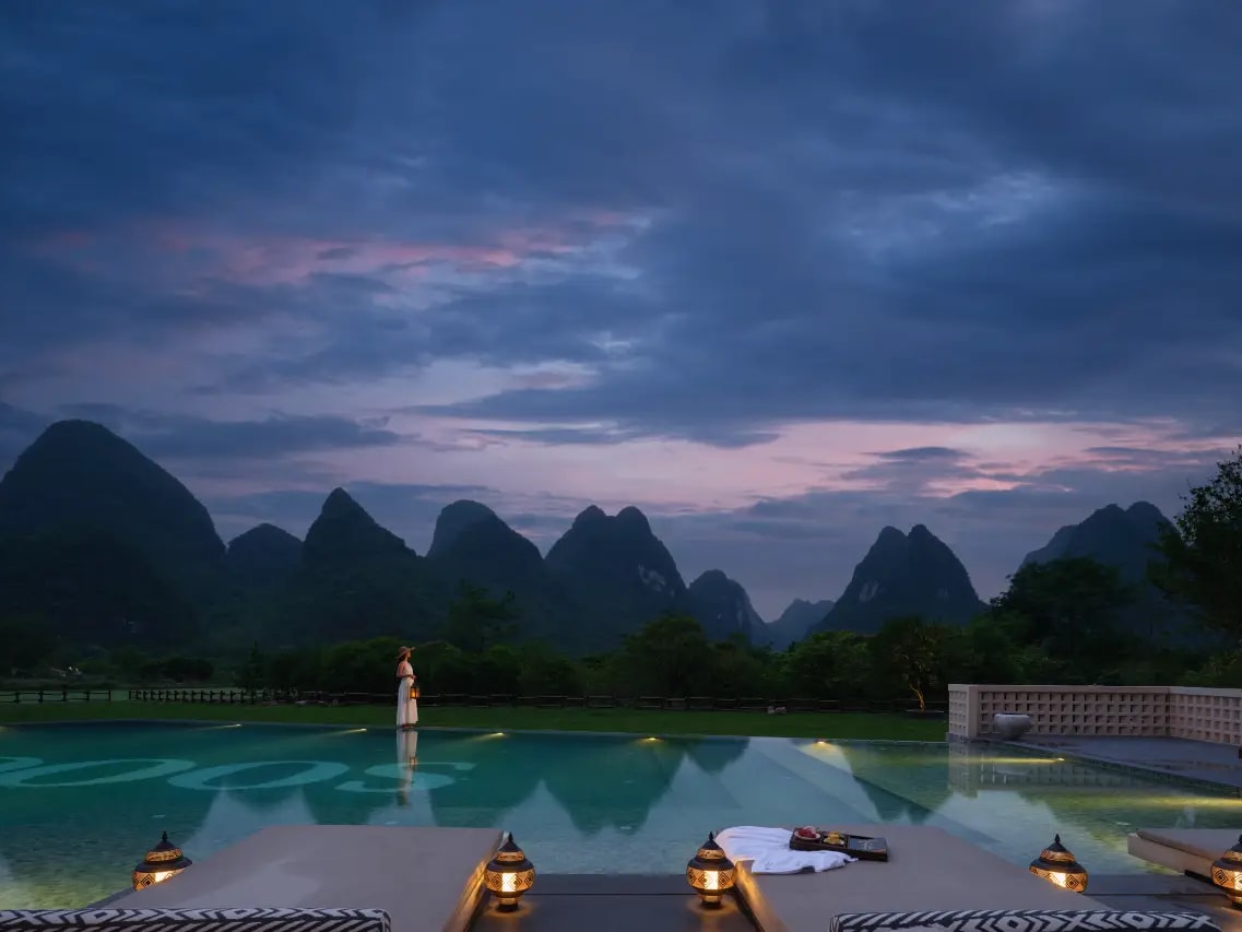Infinity pool lit at dusk with lanterns on daybeds, row of karst peaks on the horizon