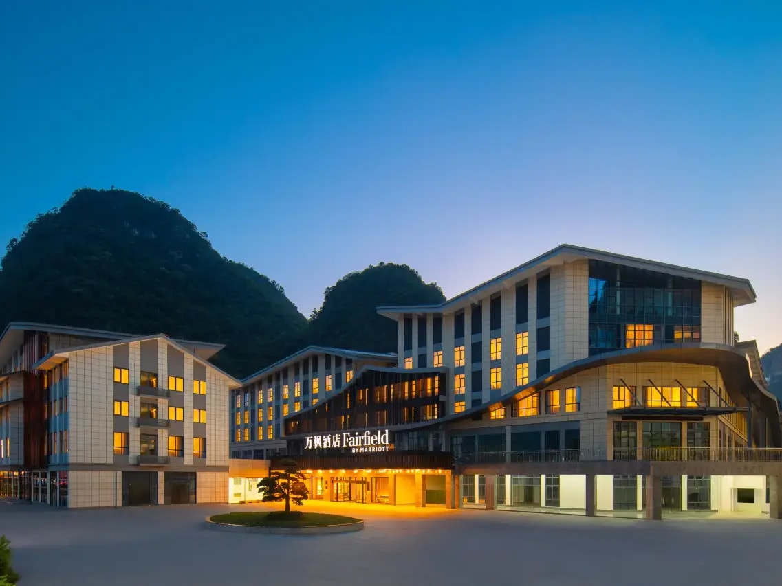 Hotel entrance lit at dusk under a Fairfield by Marriott sign, wooded karst hill behind