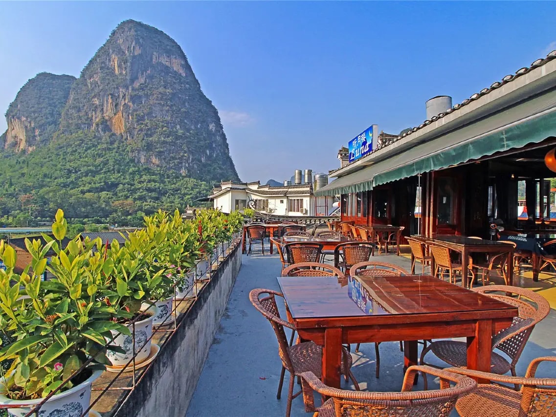Rooftop terrace of wicker chairs and wooden tables under a Luna sign, Moon Hill karst peak opposite