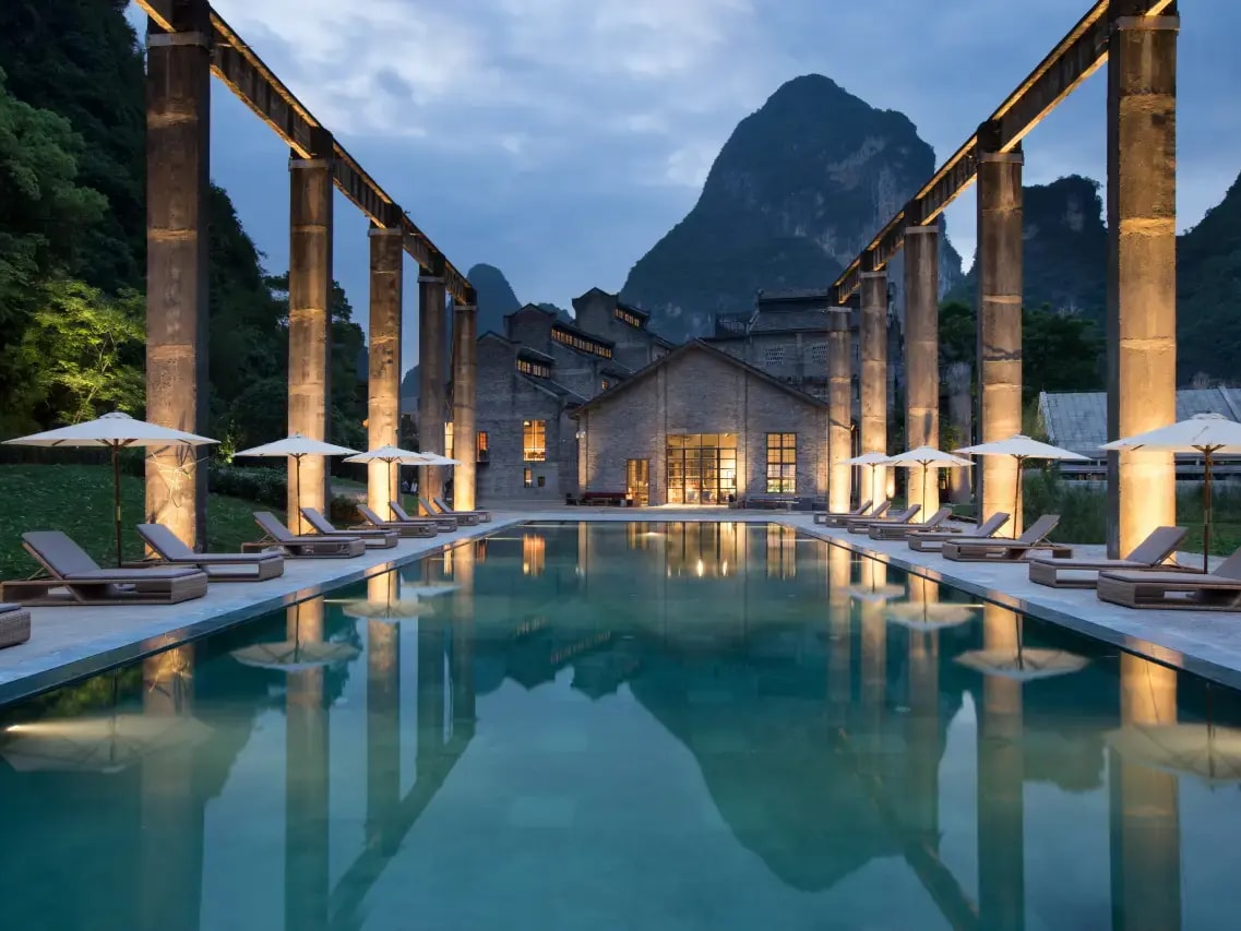 Long pool at dusk flanked by lit concrete trusses of the old mill, karst peak behind