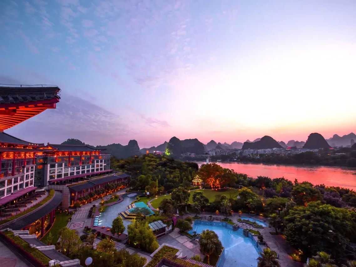 Red-columned hotel wing above lit gardens and two pools, the Li River and karst skyline at sunset