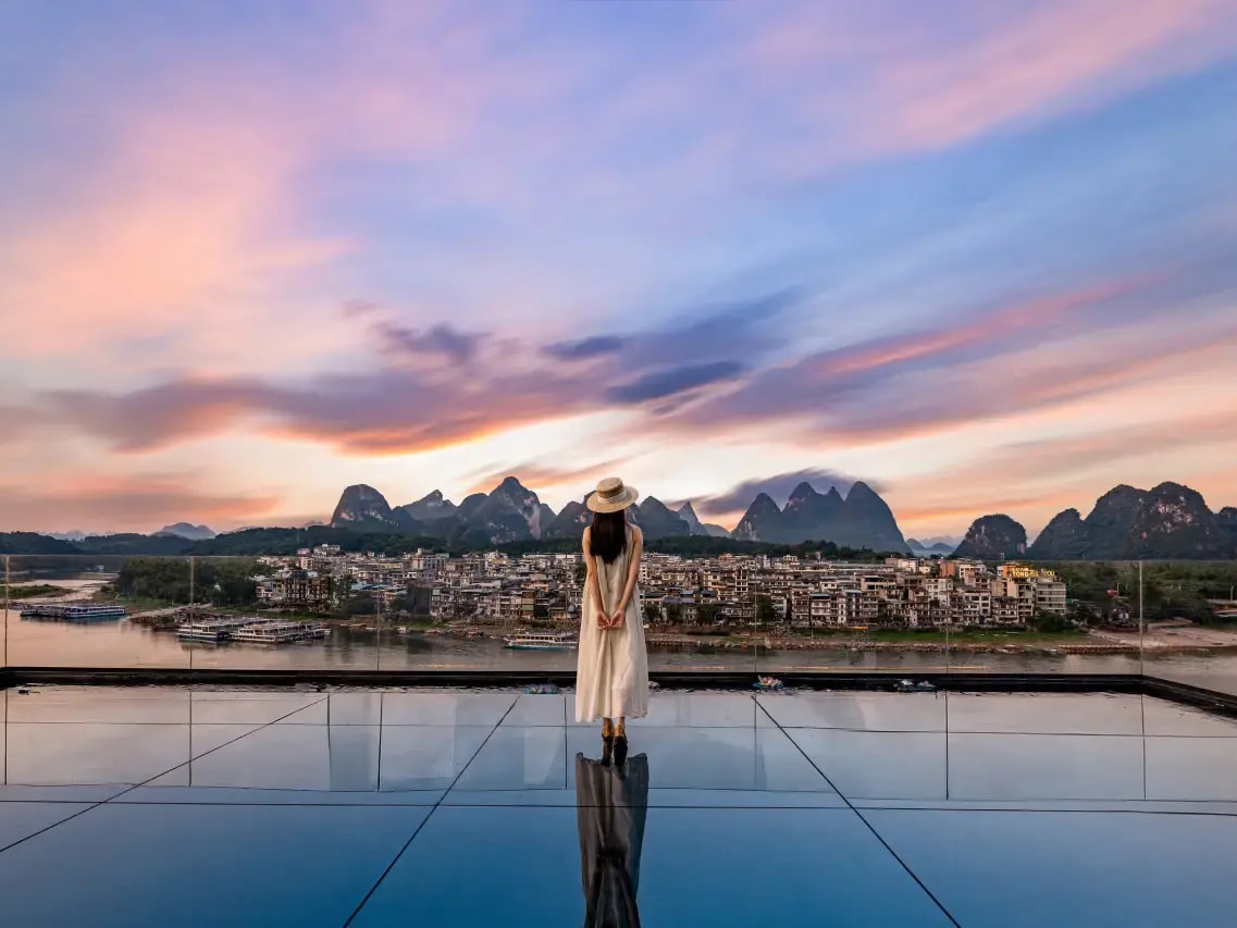 Person on a mirror-floor rooftop terrace at sunset above the Li River and the town opposite