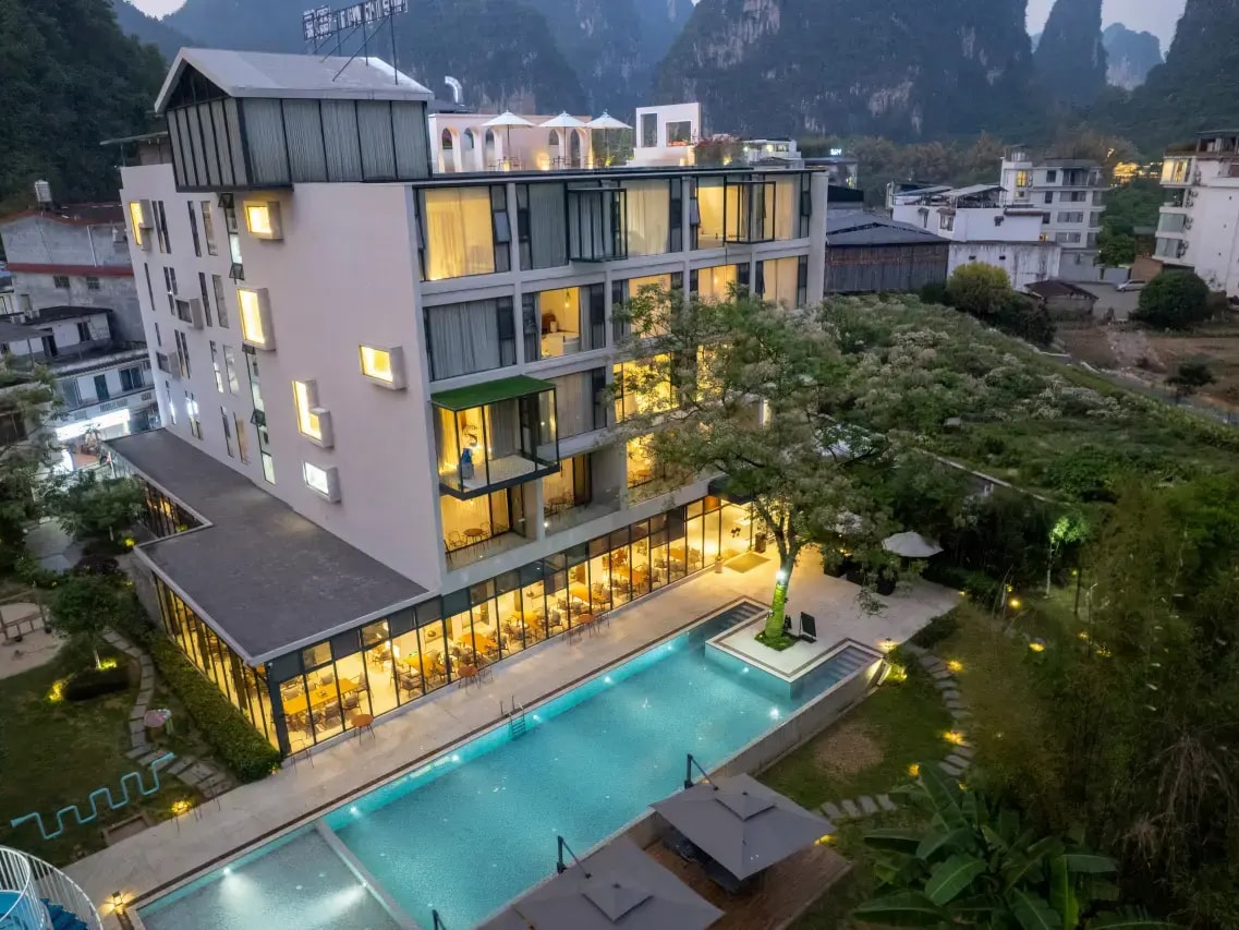 Lit hotel block at dusk above a pool and terrace, karst peaks and village behind