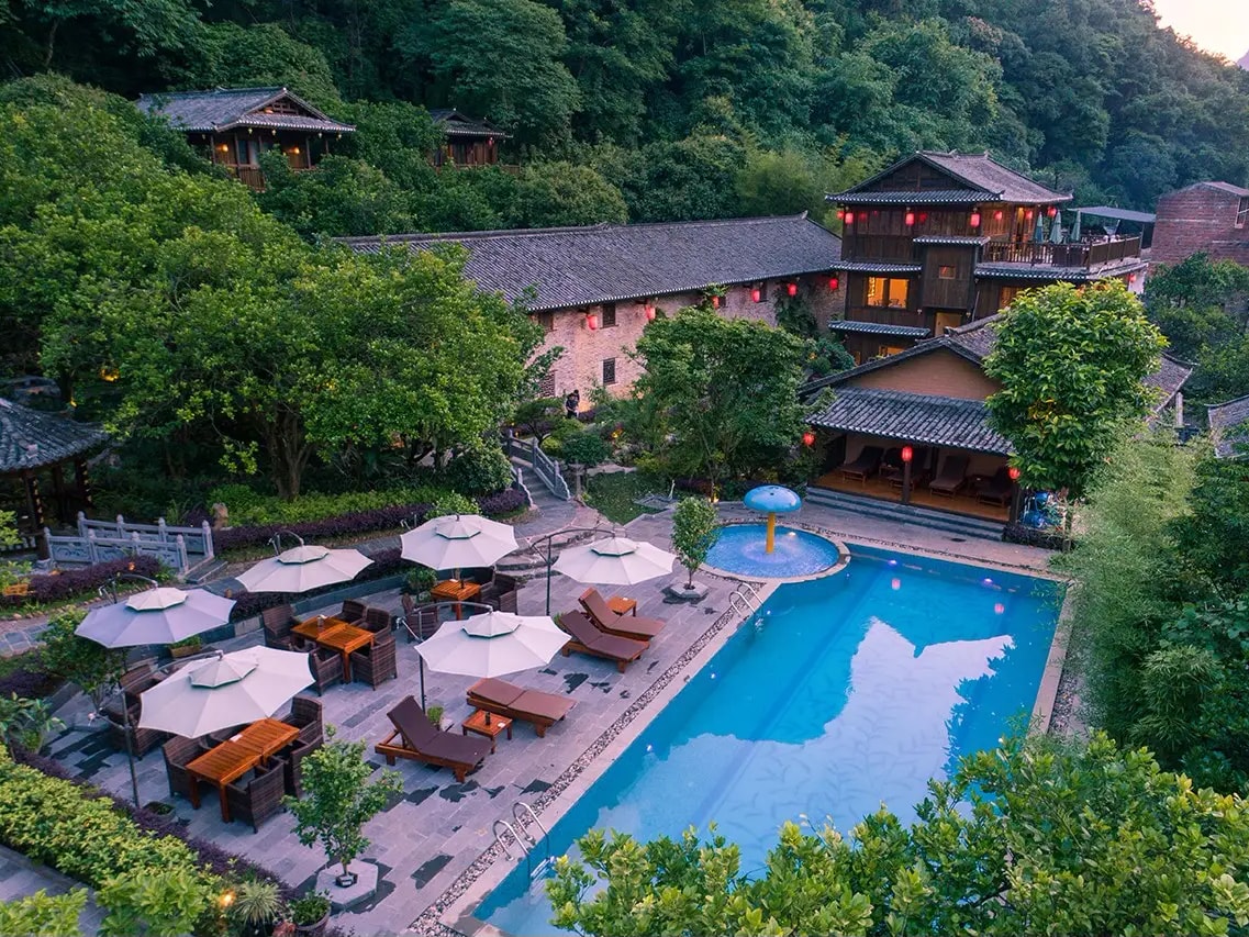 Grey-tiled traditional buildings with red lanterns above a blue pool ringed by parasols and loungers