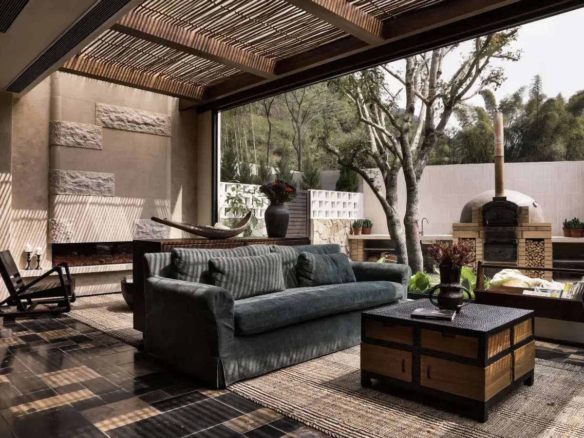 Open-sided lounge with velvet sofa and slatted timber ceiling looking onto a courtyard with a wood-fired oven