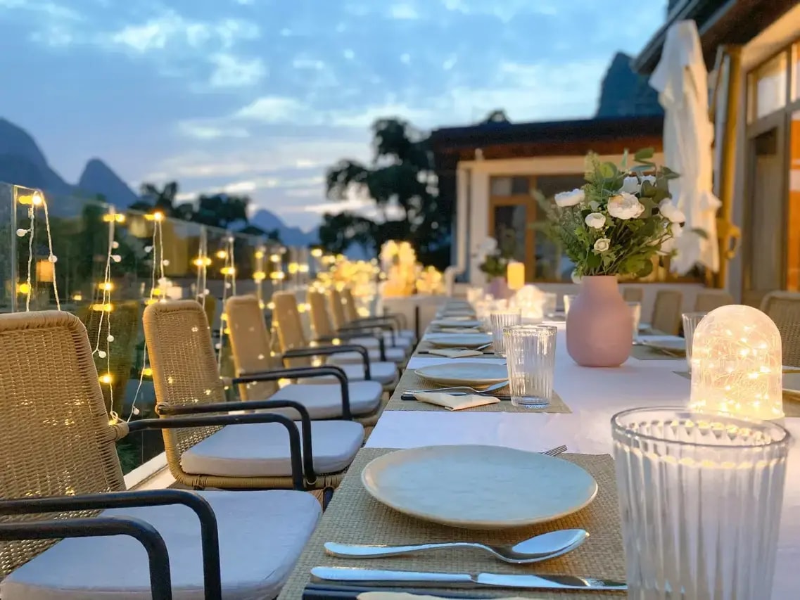 Long terrace table set for dinner under fairy lights, karst peaks fading into dusk