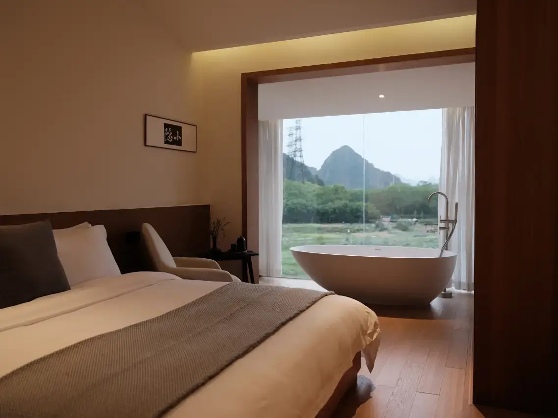 Bedroom with a freestanding tub at a full-height window looking onto a karst hill and fields