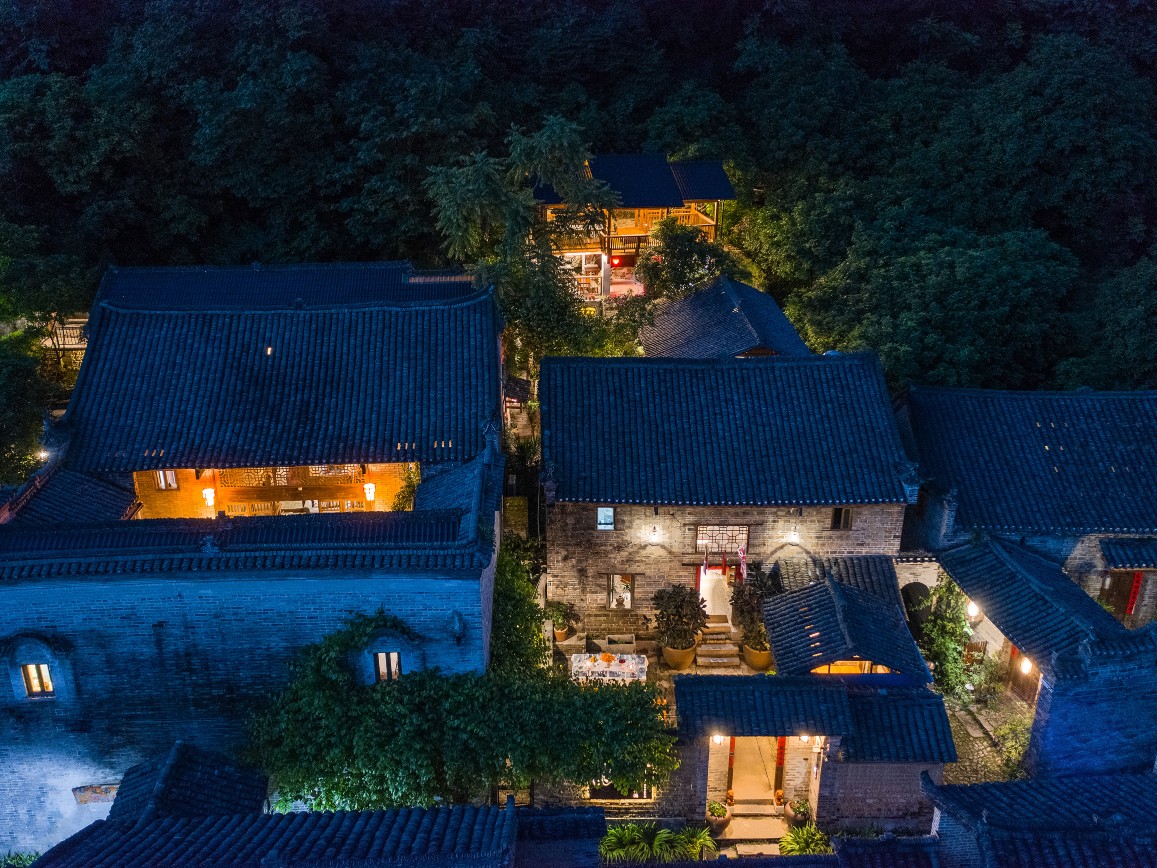 Aerial night view of grey-tiled courtyard houses lit from within, wooded hillside behind