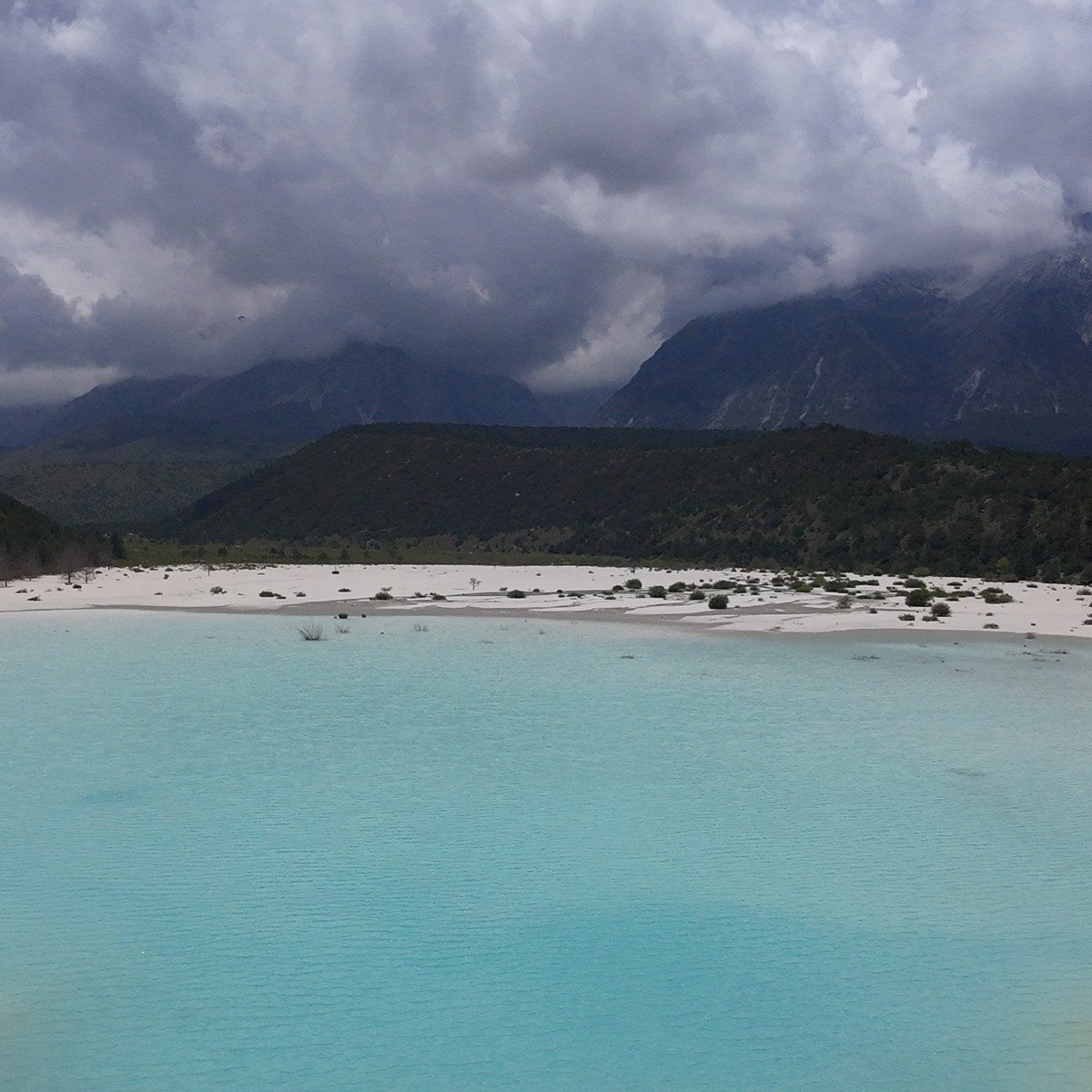 Milky turquoise lake on the white Ganheba riverbed, forested hills and cloud-capped Jade Dragon Snow Mountain behind