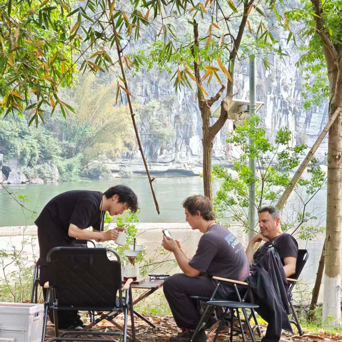 Guide brewing pour-over coffee for guests in camp chairs under bamboo by the river