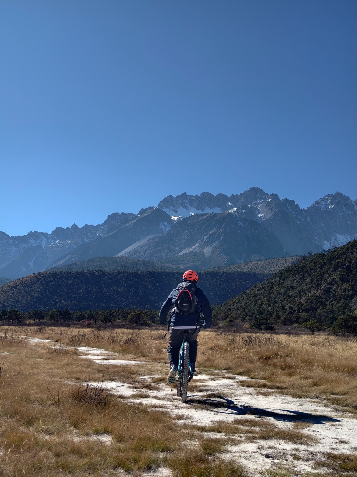 Rider on an e-MTB heading across a dry golden meadow toward the snow-streaked peaks of Jade Dragon Snow Mountain