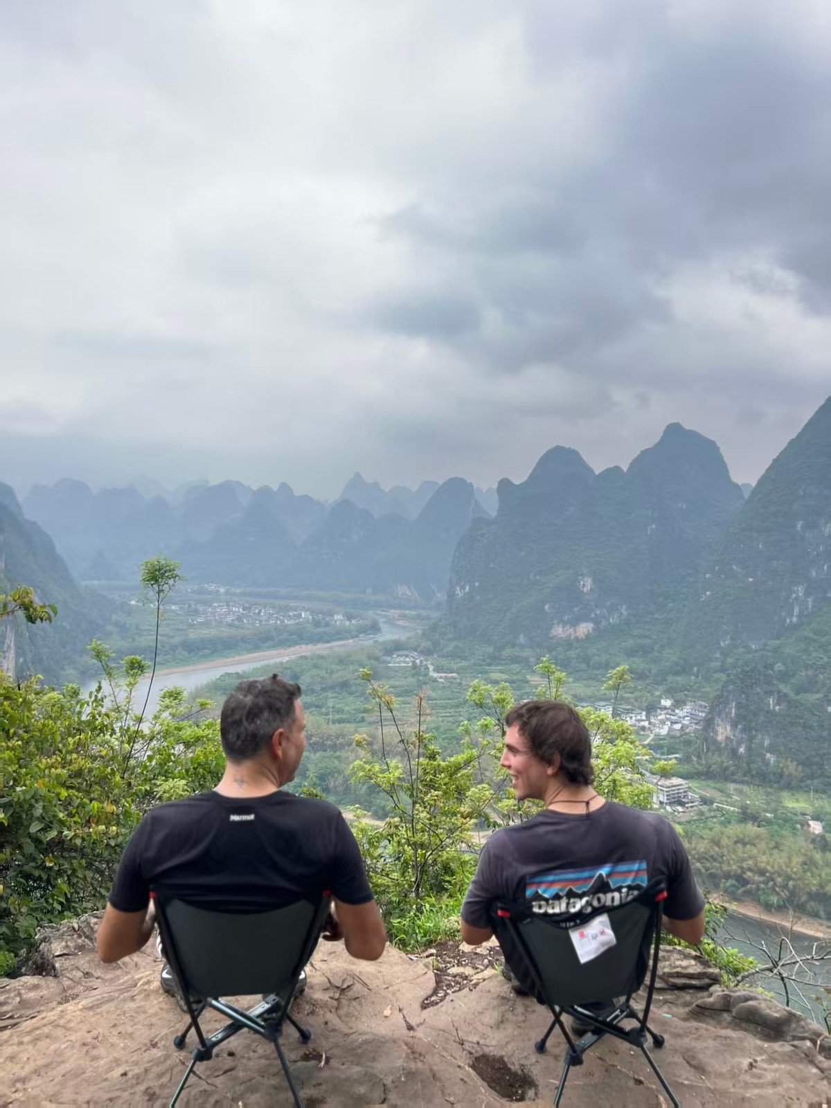 Two riders resting on camp chairs at a clifftop viewpoint over the river valley