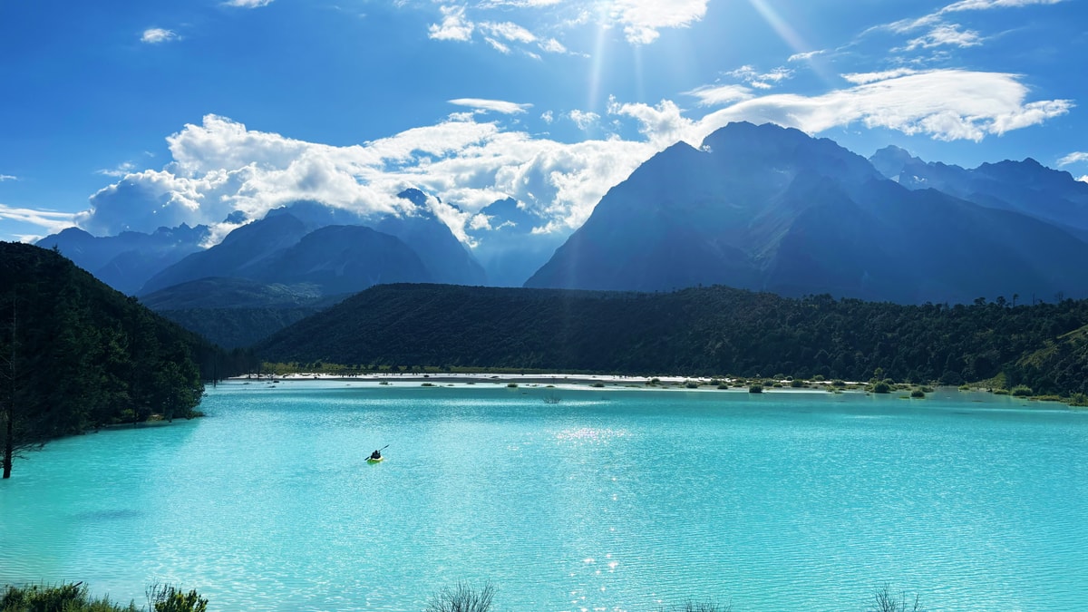 Kayaker on a turquoise lake with white shores, Jade Dragon Snow Mountain under bright sun and cloud