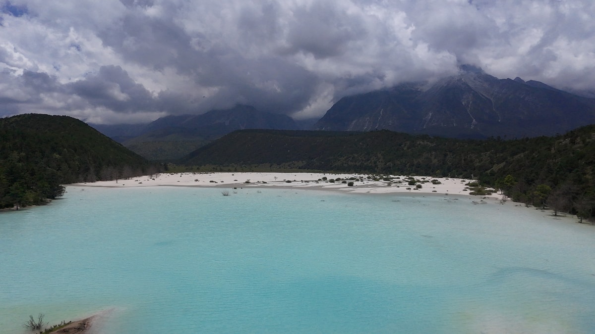 Milky turquoise lake on the white Ganheba riverbed, forested hills and cloud-capped Jade Dragon Snow Mountain behind
