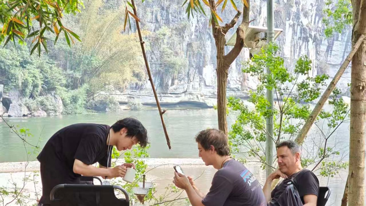 Guide brewing pour-over coffee for guests in camp chairs under bamboo by the river