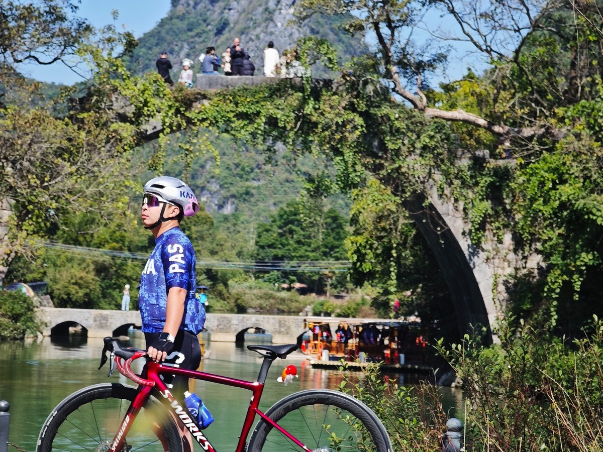 Rider with a road bike in front of an ivy-covered stone arch bridge with visitors on the deck