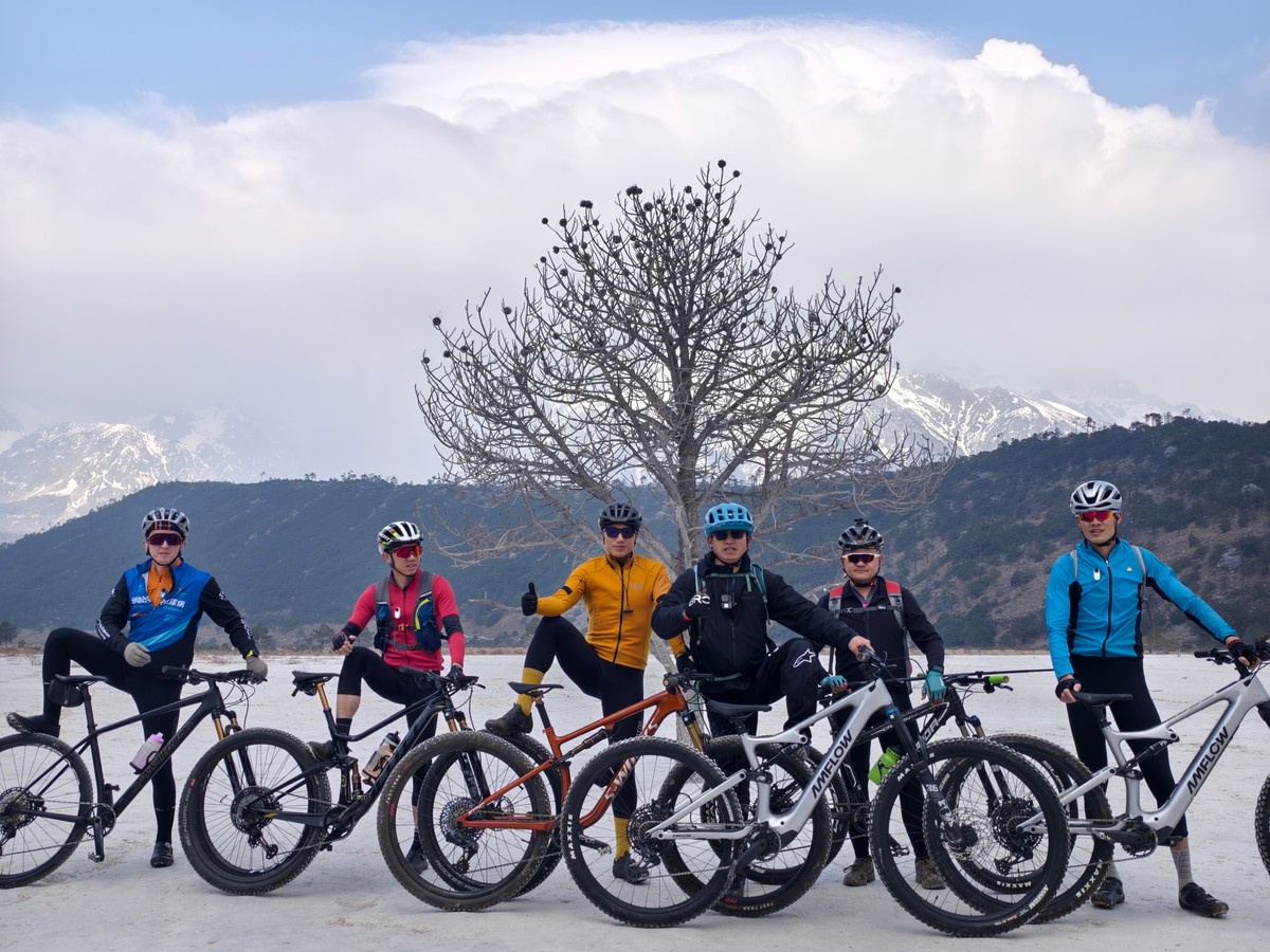 Group of riders with their bikes on the dry white riverbed under a bare tree, snow on the peaks behind