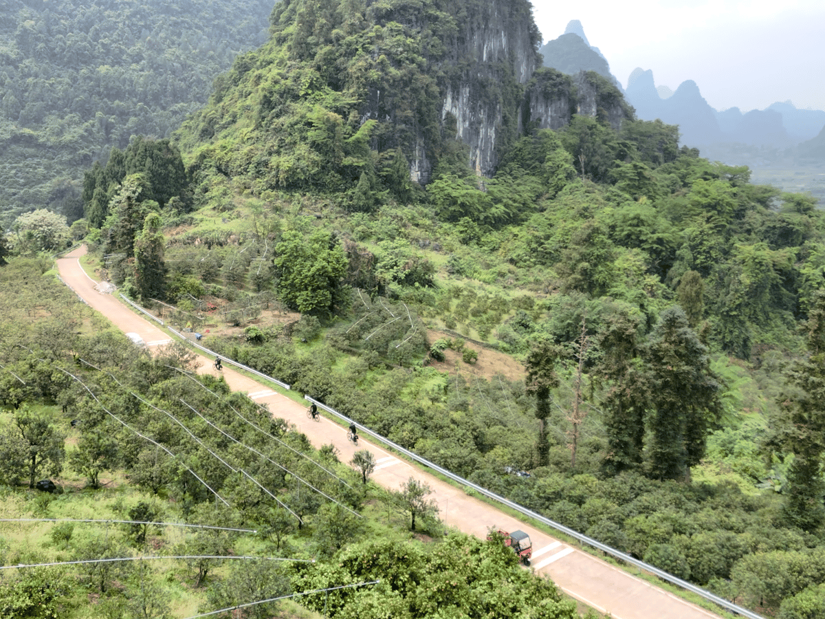 Aerial view of riders on a rural road below a limestone cliff, a three-wheeler ahead of them