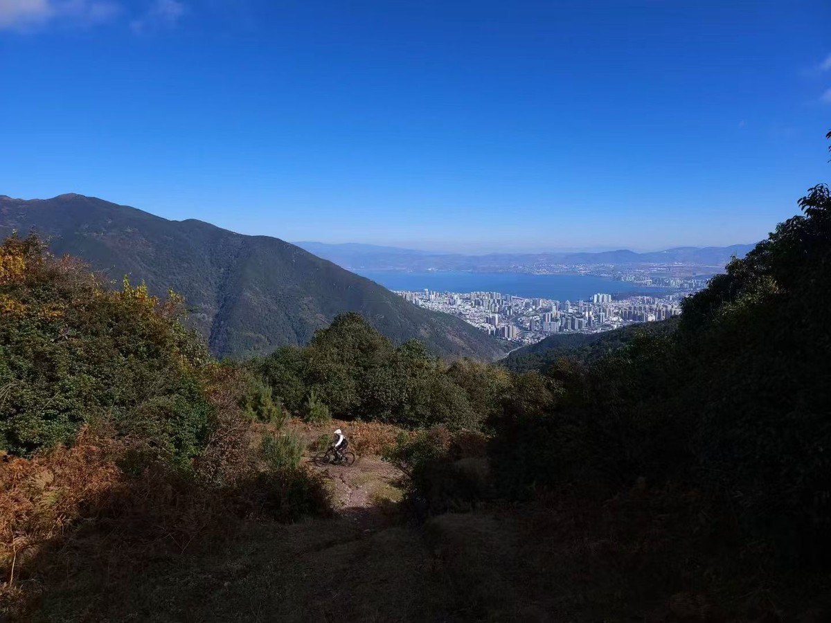 Rider on a dirt trail dropping through autumn scrub, Dali city and Erhai Lake far below