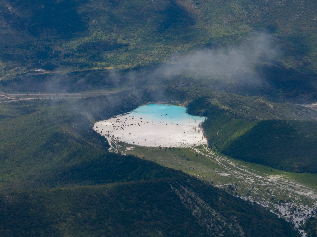 Aerial view of a turquoise alpine lake on a white sand basin, ringed by dark forested ridges and drifting mist
