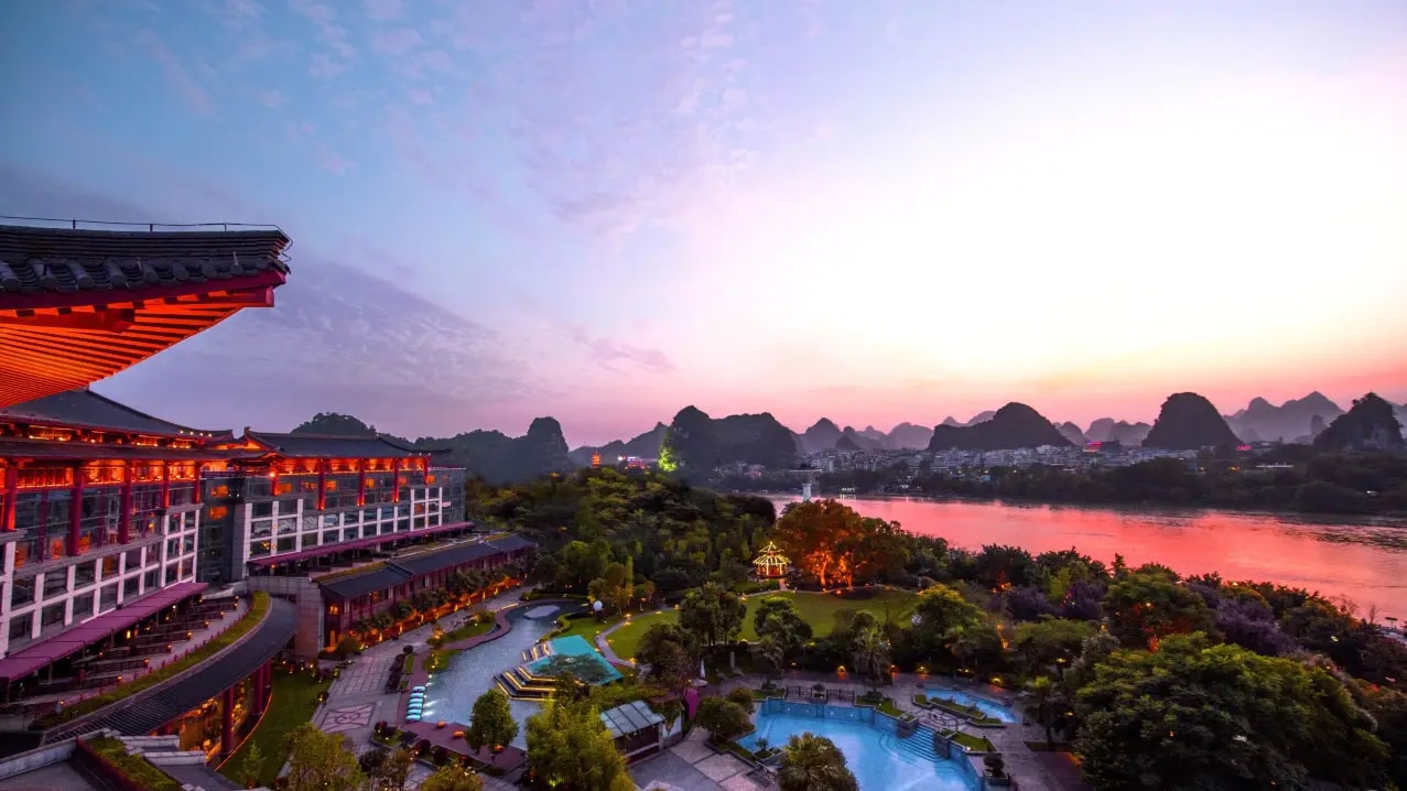 Red-columned hotel wing above lit gardens and two pools, the Li River and karst skyline at sunset