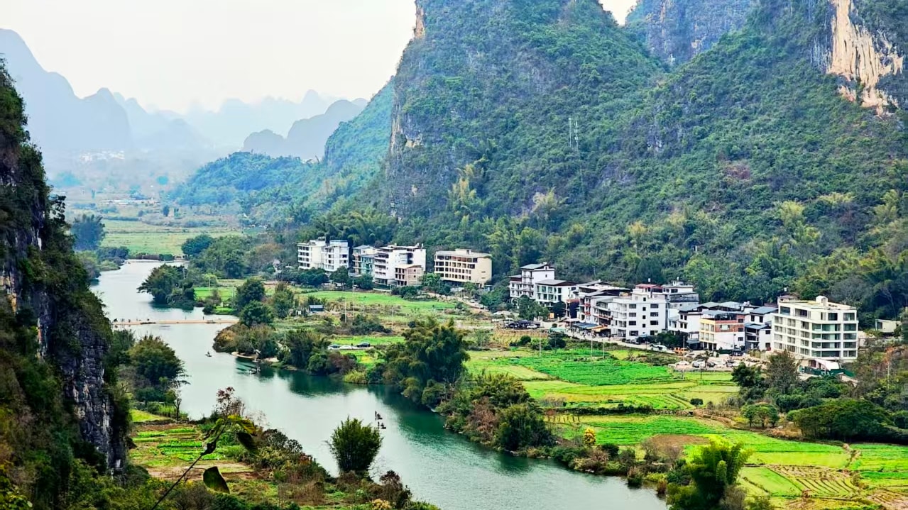 Green river below overhanging trees with a village and hills on the far bank