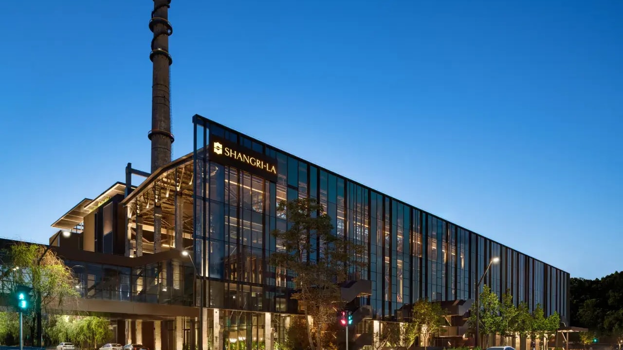 The modern glass exterior of the Shangri-La Shougang Park hotel in Beijing at twilight with industrial chimney.