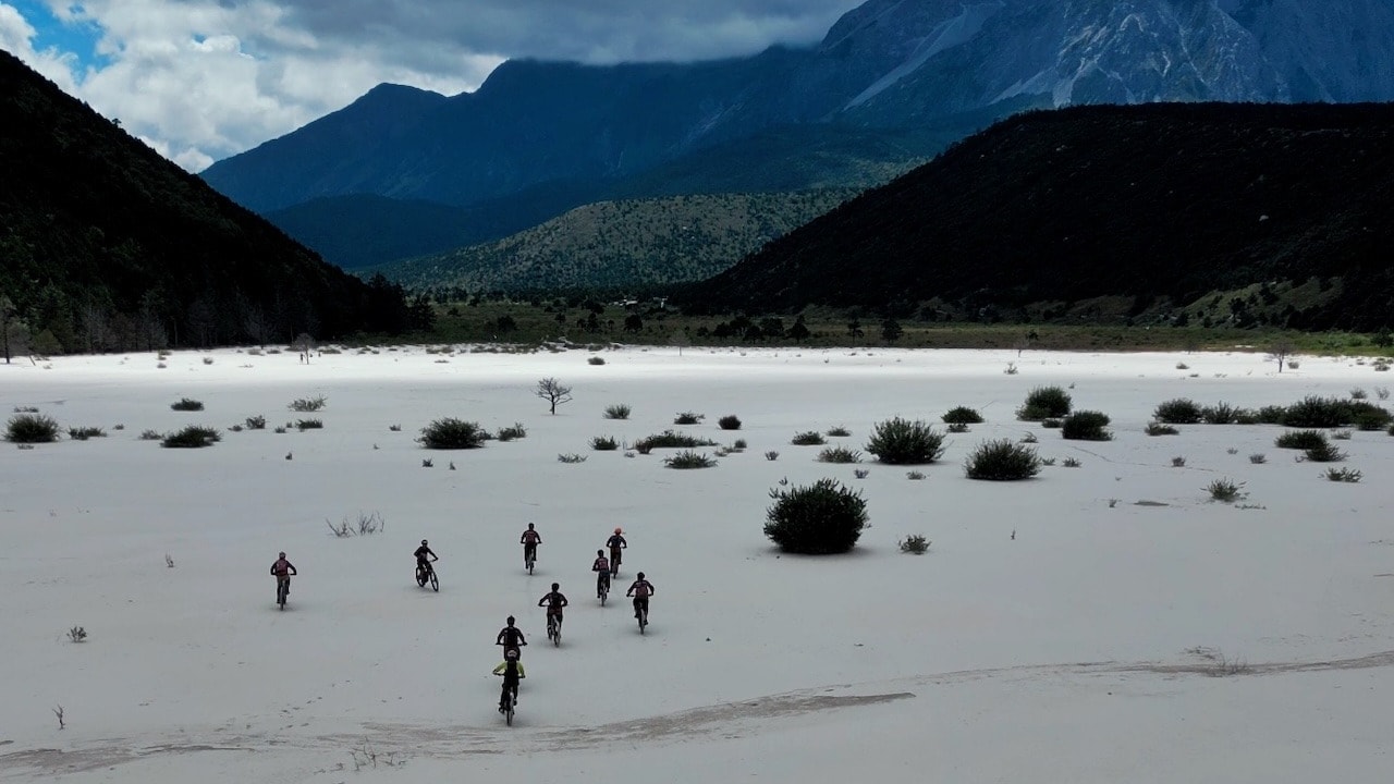 Cyclists ride across a wide white sand riverbed toward cloud-covered mountains.