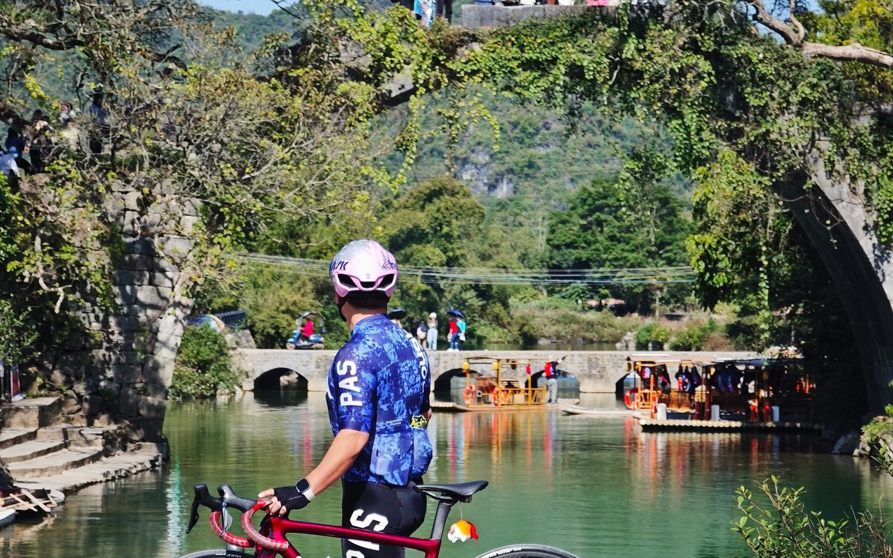 Rider with a road bike on the riverside path below an ivy-covered Ming-era stone arch bridge on the Yulong River, a karst peak behind