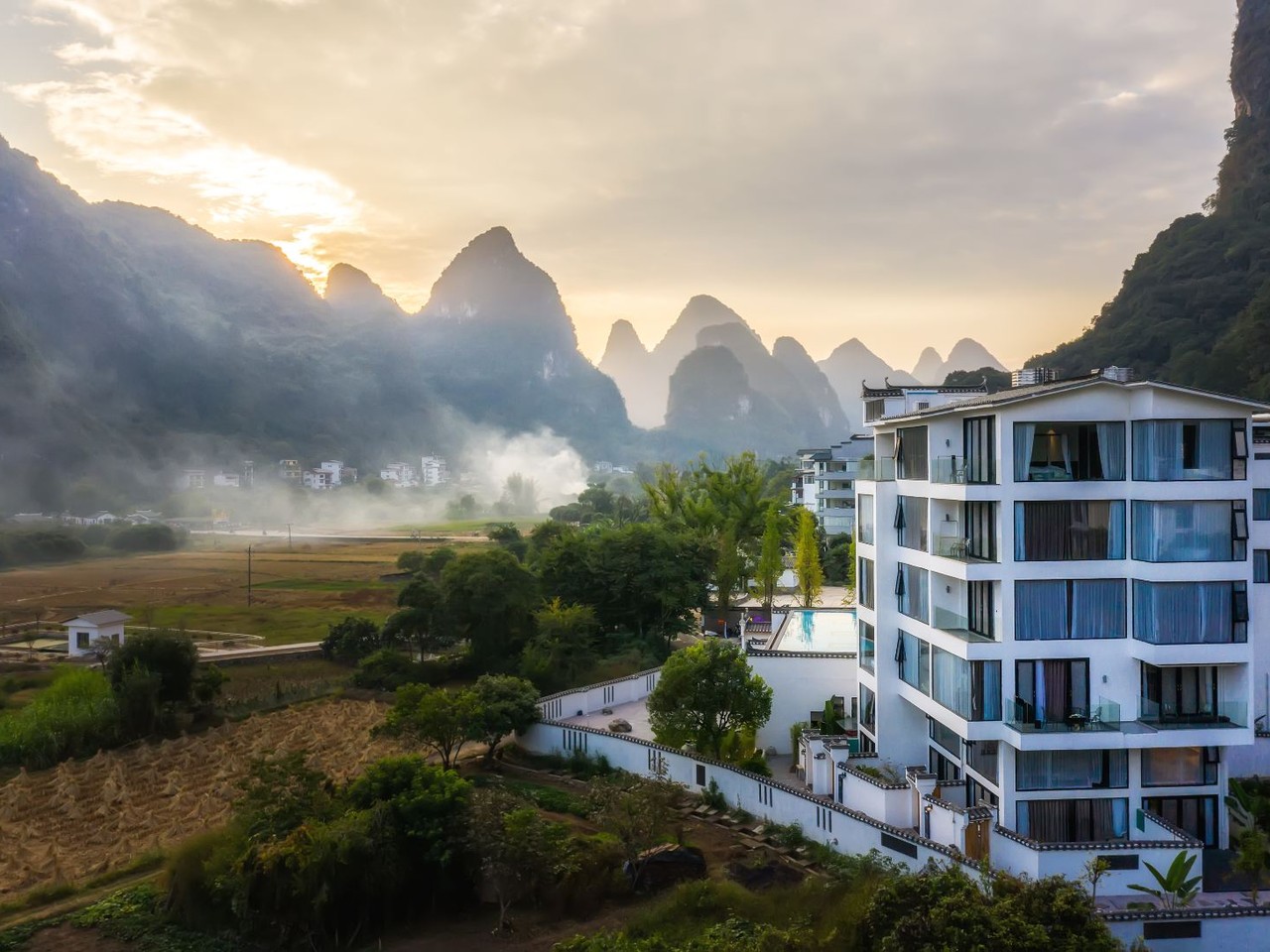 White balconied building on a hillside above mist-filled fields and karst peaks at dawn
