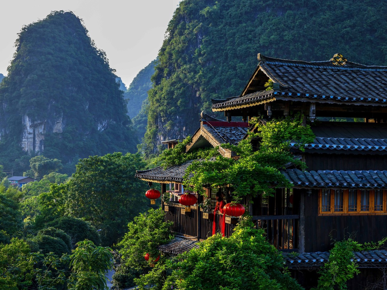 Timber and grey-tile guesthouse hung with red lanterns among trees, karst peaks behind