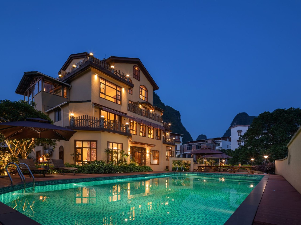 Lit three-storey villa reflected in a turquoise pool at dusk, karst peaks behind