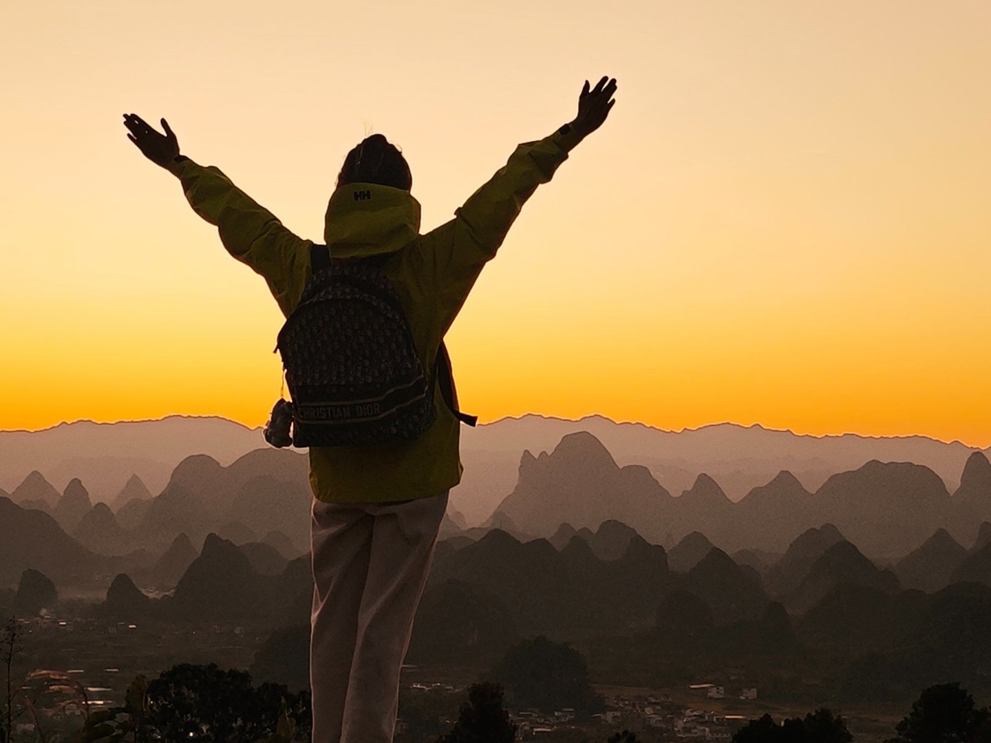 Rider with arms raised on a summit rock as the sun sets behind rows of karst peaks