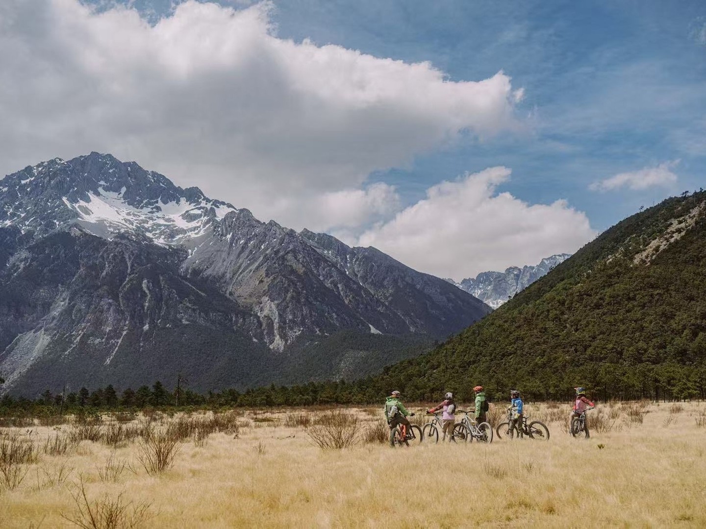 A group of mountain bikers pauses in a golden dry-grass meadow beneath the snow-streaked ridges of Jade Dragon Snow Mountain.