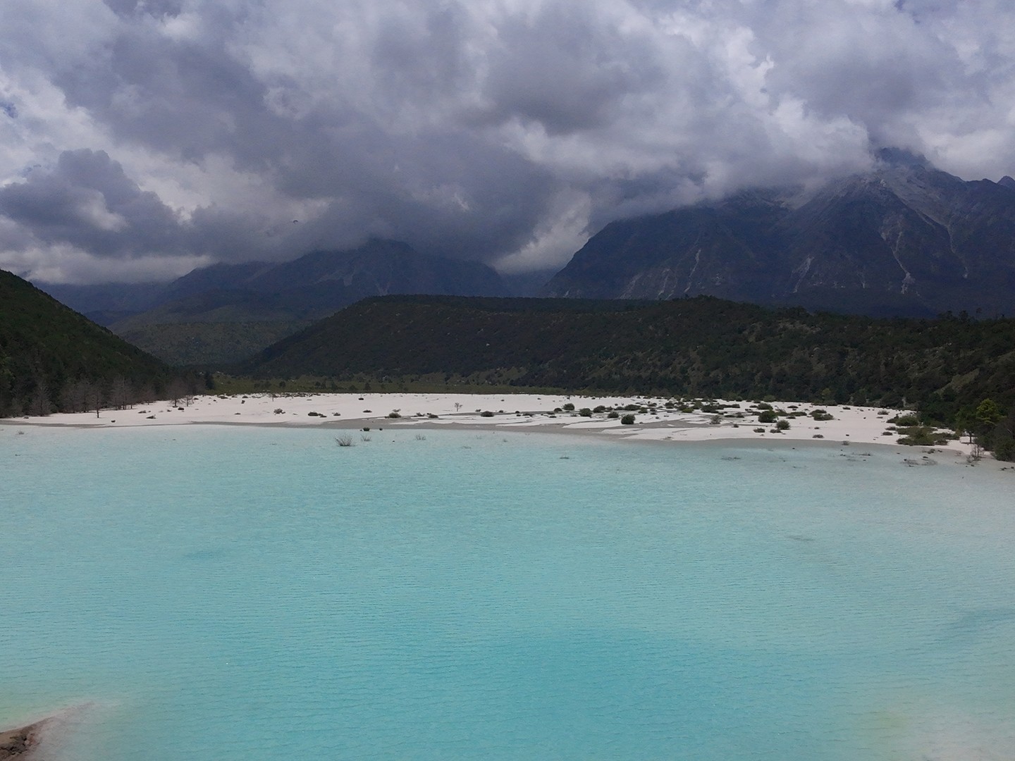 Milky turquoise lake on the white Ganheba riverbed, forested hills and cloud-capped Jade Dragon Snow Mountain behind