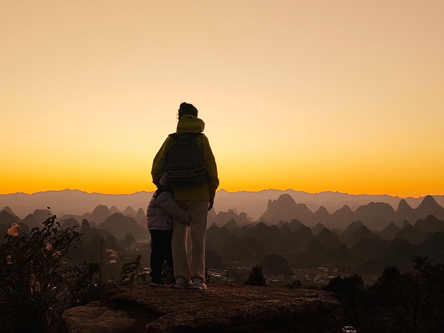 Adult and child on a summit rock silhouetted against an orange sunset over the peaks